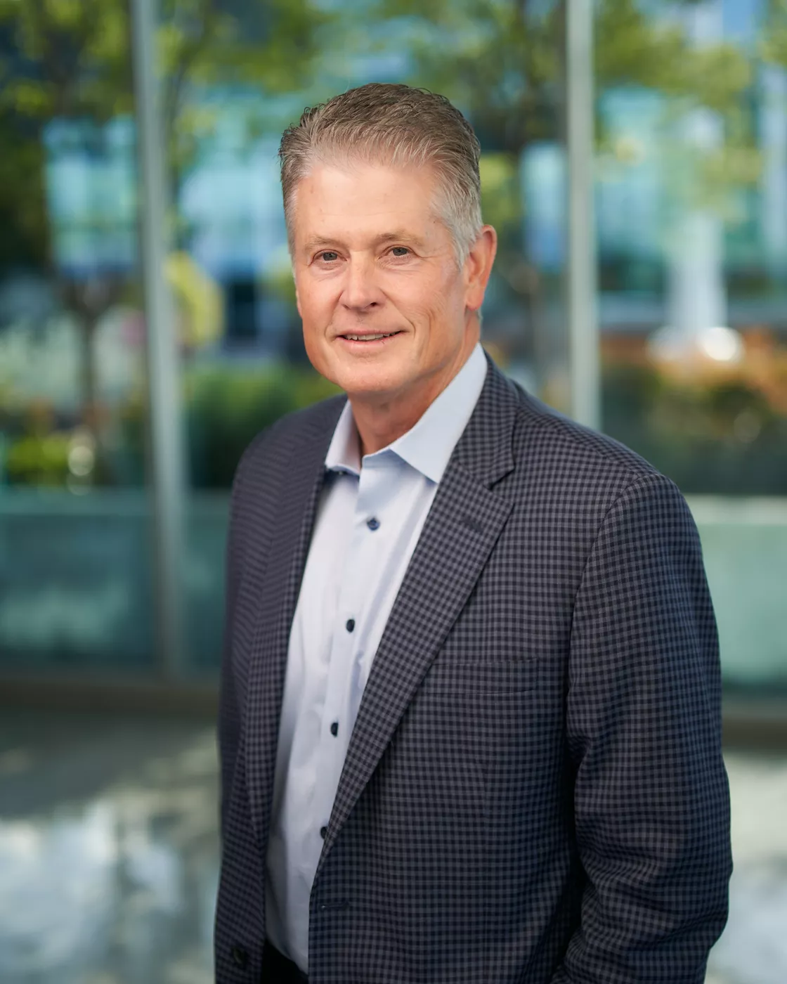 A man dressed in a checkered blazer and white shirt stands in a modern office environment. The background features large glass windows with greenery and urban elements visible outside. The setting conveys a professional and corporate atmosphere, with natural lighting enhancing the scene.